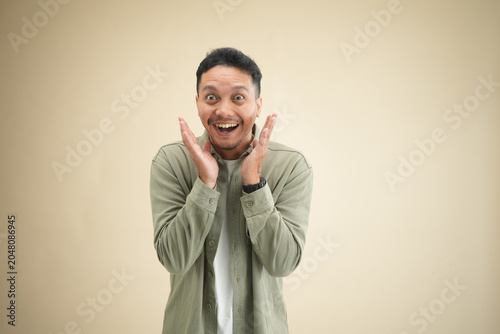Astonished young Indonesian man with hands raised near face and wide open expression against beige studio background.