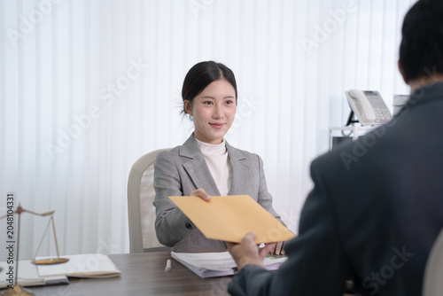 A female lawyer in a grey suit provides a yellow legal envelope to a client sitting opposite her at the office desk.