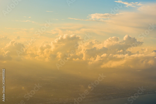 Konstfotografi Massive cumulus cloud formations illuminated by warm golden sunlight floating above a hazy distant coastal city landscape