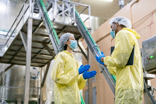 Two workers wear yellow protective suits and masks. They stand near a metal conveyor belt in an industrial facility. One gestures while conversing with another