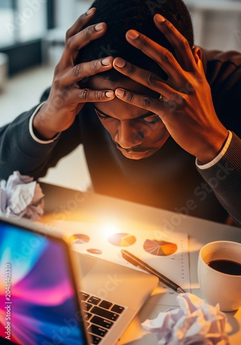 Close up of dark skin hands pressing against a forehead at a messy desk with a glowing laptop, capturing heavy stress and office pressure, light, tension, tired
