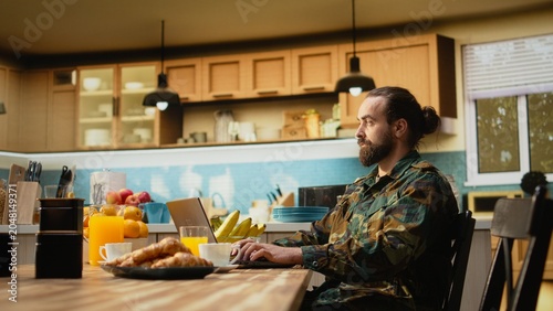Cuadro en lienzo Off duty military man, in camouflage uniform relaxing, sunlit slow morning, sitting with laptop and coffee cup and enjoying calm domestic life, Everyday routine and lifestyle away from base duties
