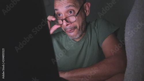 Middle-aged man with glasses getting surprised and happy while watching something on his laptop at night