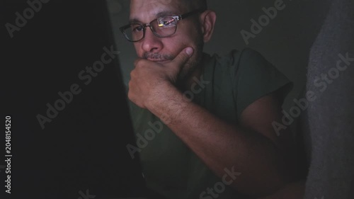 Mature man with glasses thinking and working on his computer in a dark room at home