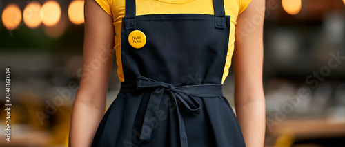 A close-up shot of a person wearing a dark blue apron tied with a bow at the waist, paired with a bright yellow t-shirt. A circular yellow badge is pinned to the apron