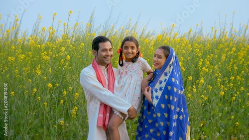 A happy rural couple lovingly holding their little daughter in their arms. Medium shot of an Indian village family happily standing in front of mustard (Sarso) agricultural field - village scene