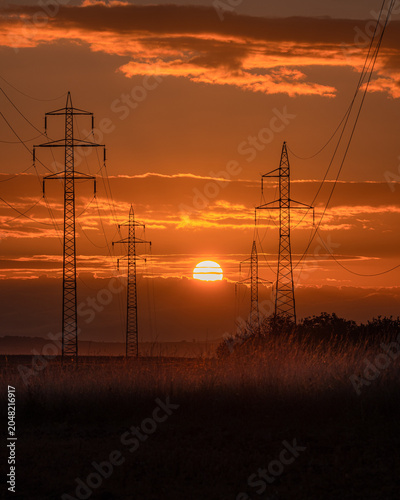 View of high voltage power lines and electricity pylons silhouetted against a vibrant orange sunset sky with clouds over a rural field in Kyjov, South Moravian Region, Czechia.