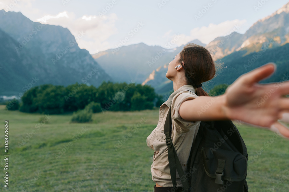 Naklejka premium Young woman with backpack stands in a sunlit meadow with distant mountains, arms outstretched as if embracing a moment of carefree outdoor freedom.