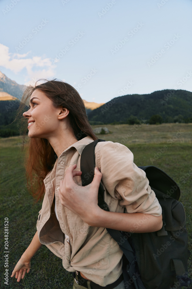 Naklejka premium A young woman with a backpack hikes through a grassy meadow with distant mountains, smiling at the peaceful landscape during golden hour.