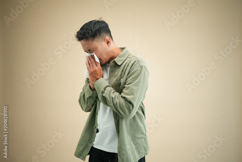 Fotografie Sick young Asian man in olive green shirt blowing nose into tissue with eyes closed against beige background