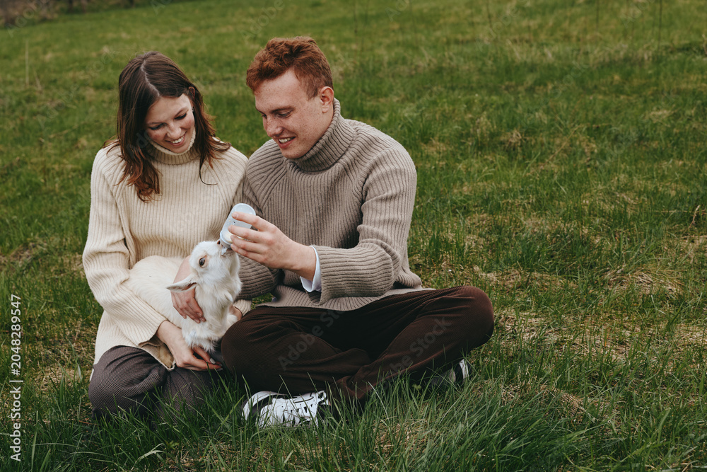 Naklejka premium Lifestyle scene with a couple feeding a goat kid outdoors on green grass, sharing affection and smiles in a bright daytime setting, portraying warm family moments outside.