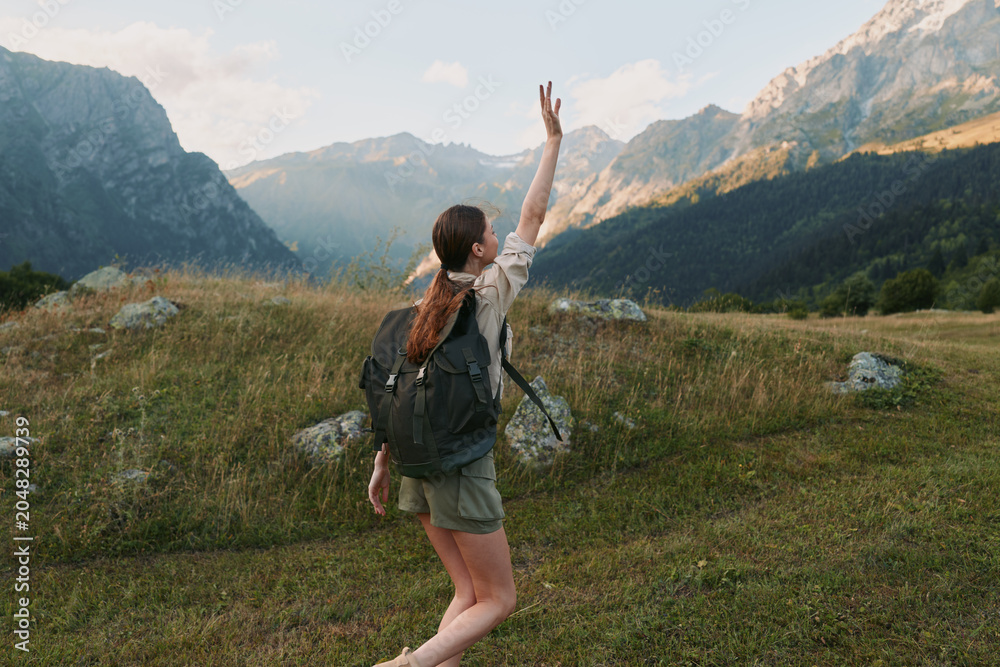 Fototapeta premium Young woman hiking outdoors in mountains, wearing backpack and casual clothes, raising hand, enjoying nature, sunny day, green grass and rocky peaks background