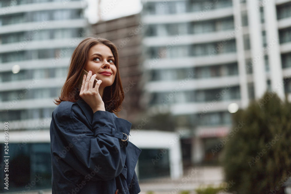 Fototapeta premium Young woman with long brown hair talks on a smartphone outdoors near modern buildings with a thoughtful expression on her face.