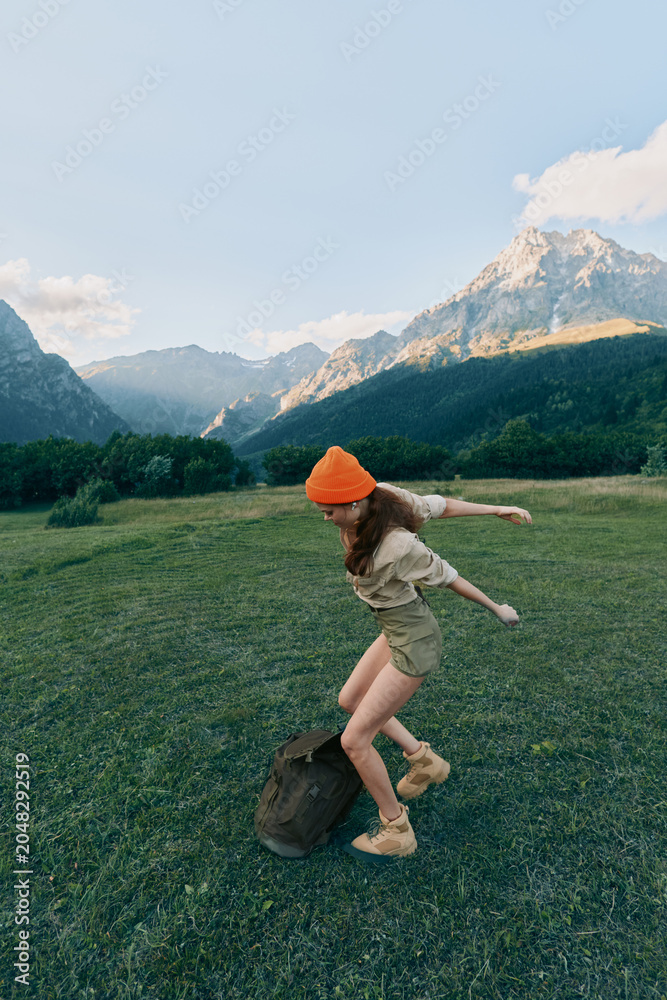 Obraz premium Young woman with long hair wearing an orange beanie jumps playfully on a backpack in a green meadow surrounded by mountains under a clear sky.