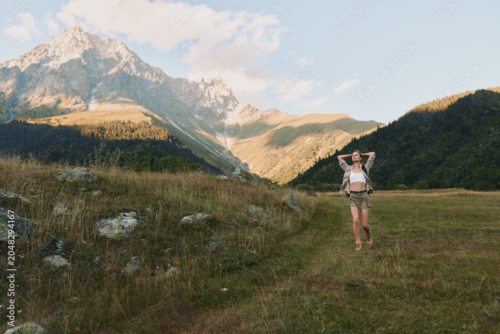 Obraz premium Young woman stands in a grassy meadow with towering mountains in the background, looking relaxed and confident.