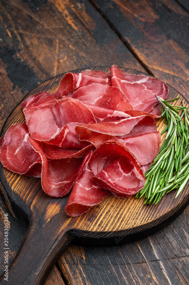 Fototapeta premium Thin cured beef slices bresaola meat arranged on a rustic wooden board with fresh rosemary over a dark textured background.