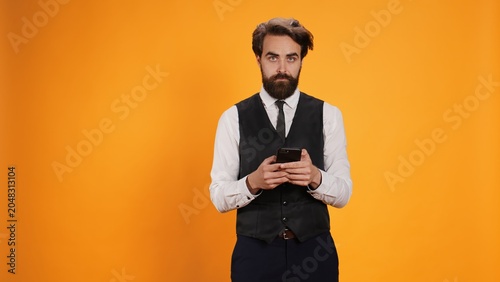 Stylish waiter uses smartphone to text before serving customers at table  browsing online pages against yellow background. Professional valet working at fine dining restaurant.