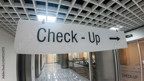 Hanging Check-Up directional sign with a right arrow in a bright, modern hospital interior corridor. Preventive medicine and healthcare wayfinding.