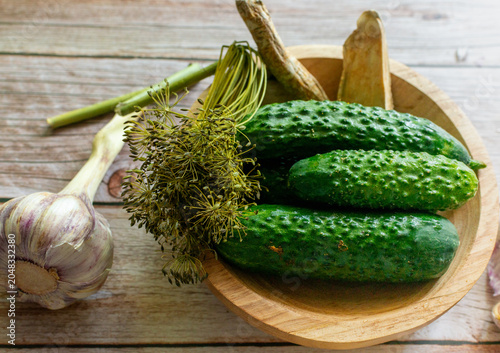 Fresh cucumbers in a wooden bowl with dill umbels, garlic and horseradish root on rustic wooden background. Useful for pickling, home cooking, fermentation and vegetable harvest concepts.