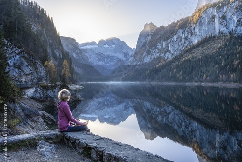 A woman doing yoga by the lake. Lotus position (Padmasana) . Vorderer Gosausee in autumn with a view of the Dachstein mountains. The Gosaukamm on the right. Blue sky, good weather. Reflection. Morning atmosphere. Vorderer Gosausee, Gosau, Gosau valley, Salzkammergut, Upper Austria, Austria