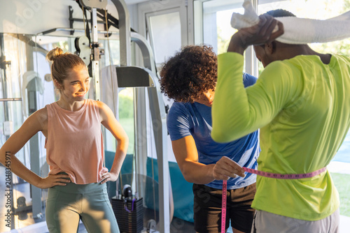 Fotografía Diverse trio doing waist check in gym, trainer wrapping pink tape, client holdin