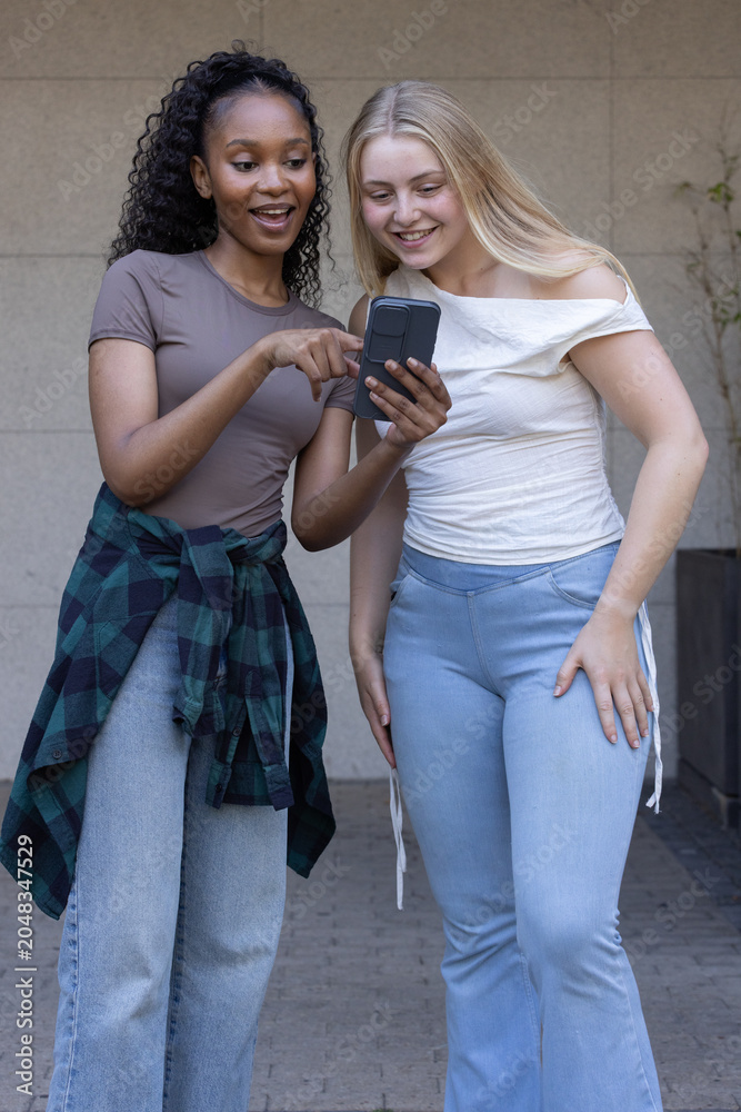 Naklejka premium Diverse female friends standing close, sharing phone, one wearing plaid shirt at waist in courtyard