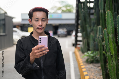 Foto Non-binary gay Thai man from Isan holding smartphone in an urban street setting, showcasing authentic gender fluid identity, nonconforming pride, and queer representation