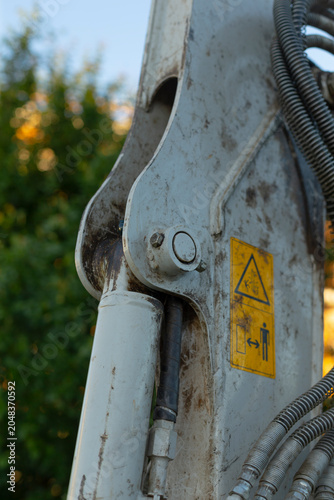 Detailed close-up of a heavy construction machine arm showing a worn metal joint, hydraulic cylinder, flexible hoses, and a yellow safety warning label