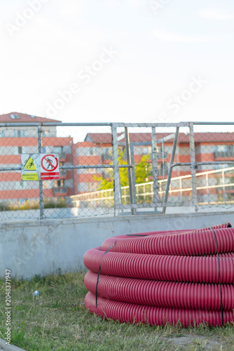 A large coiled pink corrugated pipe rests on grass beside a concrete wall and metal fence, with warning signs and residential buildings in the background