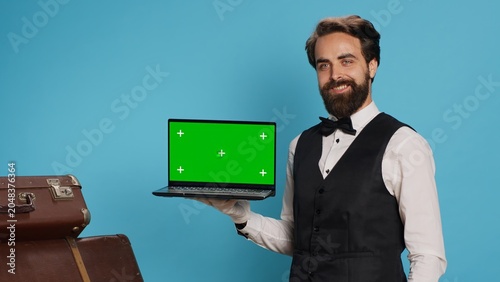 Photos Classy bellboy shows greenscreen on laptop in studio while he wears white gloves next to guests suitcase