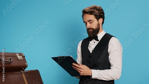 Tableau sur toile Bellboy reading checklist of guests, holding ledger or register on clipboard to verify list of travellers arriving at hotel
