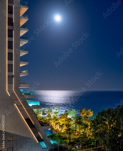  balconies of a stylish modern high-rise building on the beach with palm trees at Limasol in the night with brilliant moonlight and a shimmering sea stretching to the horizon, Cyprus