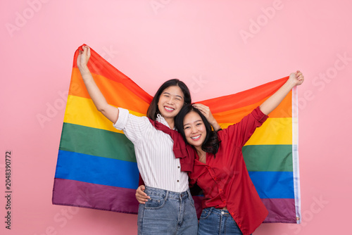 Young Asian lesbian couple holding a rainbow flag standing isolated on a pink background.