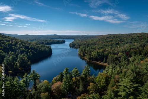 Aerial view of a winding lake surrounded by lush green and autumnal forests under a clear blue sky with whispy clouds