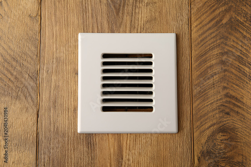 Close-up of a square, white vent cover with horizontal slots set flush into a wooden floor with prominent grain
