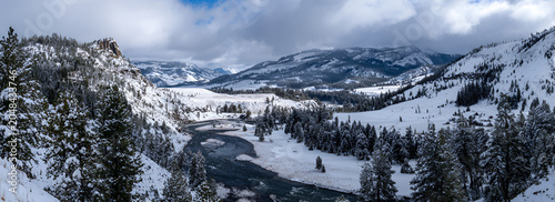 A scenic panorama of a snowy mountain range and a river winding through a valley, trees covered in white. Cloudy sky