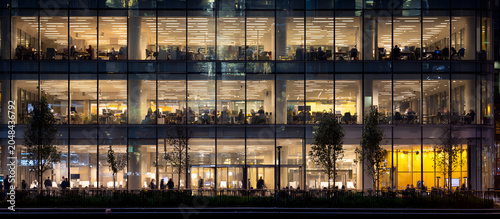 Modern office building facade at night, featuring illuminated interiors with multiple levels and reflective glass. People silhouettes
