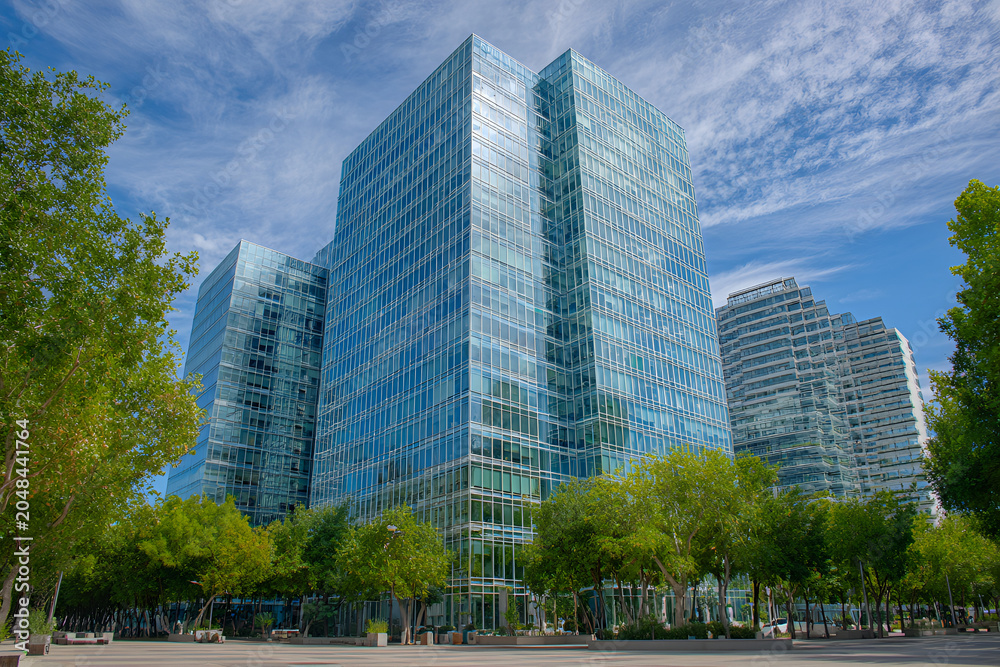 Fototapeta premium Modern glass skyscrapers rise against a blue sky with fluffy clouds, surrounded by lush green trees and a paved area