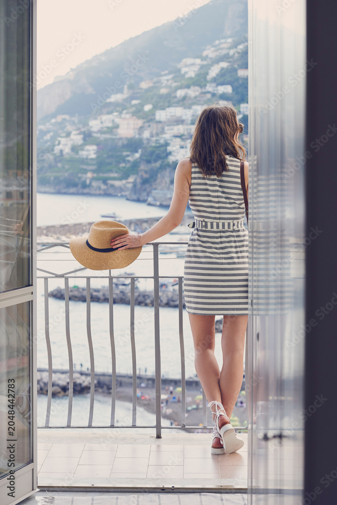 Fototapeta premium Woman, travel and ocean view from hotel with balcony, thinking and hat for summer holiday outdoor. Back, person or tourist reflection in accommodation with terrace, beach scenery or vacation in Italy