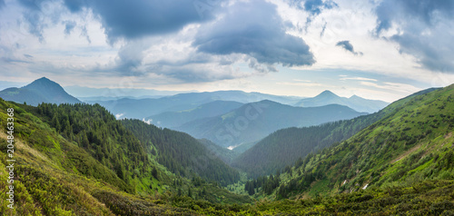 Canvas Print Panoramic view of a lush mountain valley framed by forested slopes and distant peaks beneath dramatic storm clouds, with soft light illuminating the layered landscape