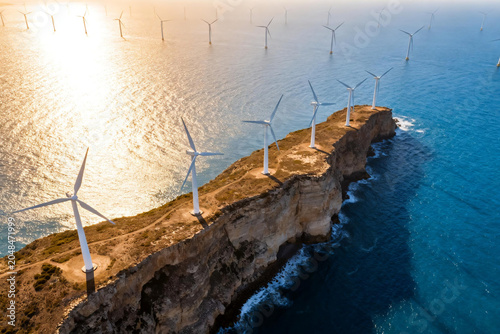 Wind turbines on dramatic cliff edge and offshore wind farm in the ocean at sunset, sustainable green energy infrastructure landscape