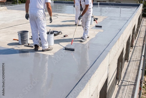 Construction workers in white overalls applying waterproofing sealant to a flat rooftop with rollers.