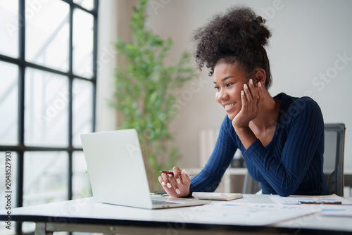 Happy young Black woman smiling, working on laptop computer at modern office desk, enjoying remote work, online learning, or video call while collaborating remotely
