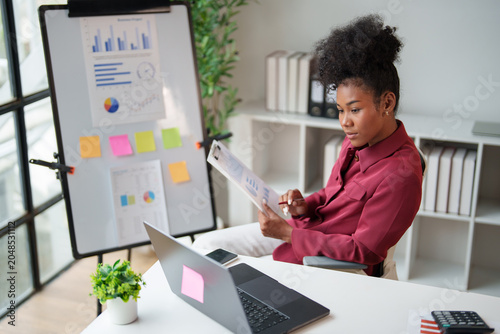 African American businesswoman analyzing financial data, reviewing statistics and business reports on documents and a laptop in a modern office for smart planning and development
