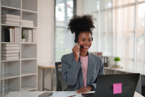 African American woman at a modern help desk wearing a headset and smiling while using a laptop to provide friendly, professional remote customer support and virtual assistance