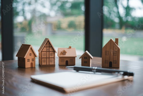 Little wooden houses arranged on table near notebook and pen, symbolizing real estate investment or housing market choices.