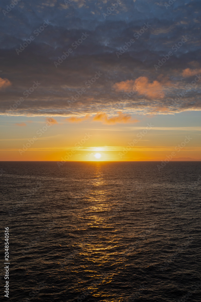 Obraz premium Golden sunset over calm ocean horizon with distant mountains and dramatic cloudy sky