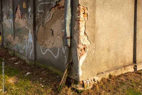 Billede på lærred Old exterior wall with cracked plaster, exposed red brick, graffiti marks and drainpipe in warm sunlight