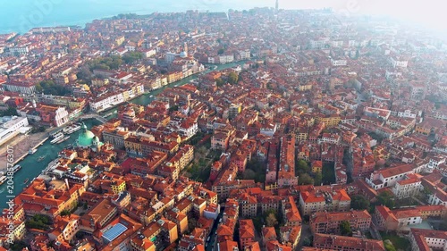 4K aerial drone shot of Venice, Italy, capturing the iconic Grand Canal winding through a dense historic cityscape of red terracotta rooftops and ancient architecture against a hazy morning horizon.