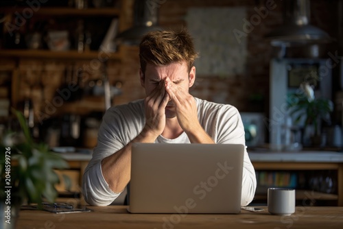 Tired young man rubbing eyes with hands sitting in front of laptop in kitchen on stressful workday